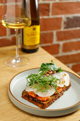 Toast with grilled shrimps and vegetables, poached egg and herbs on top served in restaurant. Wine bottle and glass on background