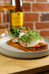 Toast with grilled shrimps and vegetables, poached egg and herbs on top served in restaurant. Wine bottle and glass on background
