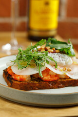 Toast with grilled shrimps and vegetables, poached egg and herbs on top served in restaurant. Wine bottle and glass on background