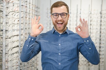 Naklejka premium In Optics Shop. Portrait of male client holding and wearing different spectacles, choosing and trying on new glasses at optical store. Man picking frame for vision correction, closeup.
