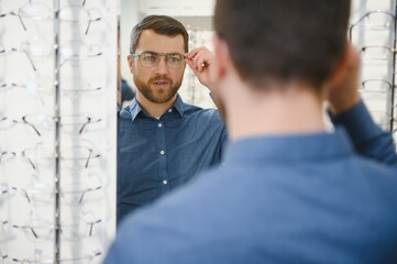 In Optics Shop. Portrait of male client holding and wearing different spectacles, choosing and trying on new glasses at optical store. Man picking frame for vision correction, closeup.