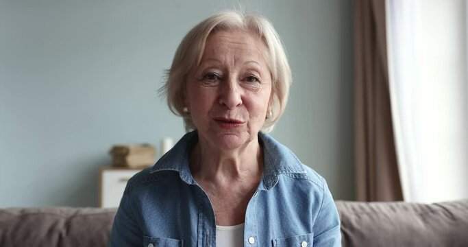 Head shot portrait smiling older woman sit on sofa making call using videoconference application, welcoming grown up children, enjoy pleasant remote comfortable communication use modern tech at home