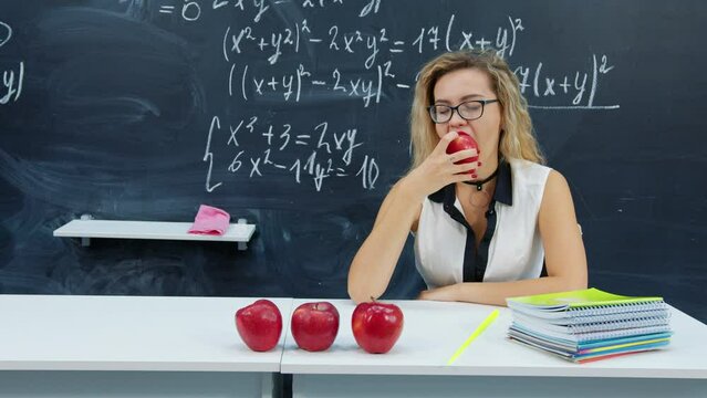 Sexy blonde girl in glasses sits at desk near blackboard, takes, bites off ripe apple. Young math female teacher eats fruit in classroom. Beautiful student makes snack at break at university