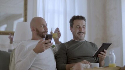 Happy gay couple having breakfast in bed and using gadgets. Cheerful homosexual men in pajamas reading news on tablet and phone, talking, spending morning together. LGBT, love, morning routine concept - Powered by Adobe