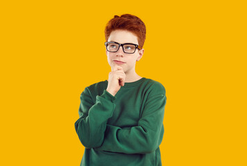 Thoughtful teenage boy standing with his hand on chin. Handsome puzzled redhead boy wearing glasses posing with serious face expression over yellow isolated background