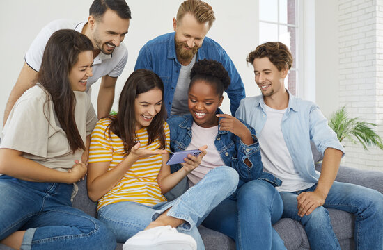 Group of people gathered together around female friend with mobile phone and are watching funny photos and videos together. Smiling multiracial millennial friends relaxing together on comfortable sofa