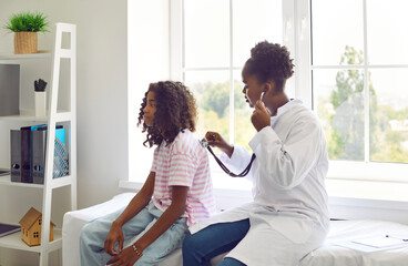Female doctor listens to back of preteen girl patient to check her lungs, breathing and heartbeat. Doctor listens with stethoscope to African American teenage girl sitting on examination couch.