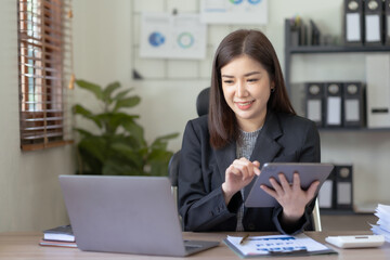 Asian business woman sitting happily with laptop and using tablet and taking intense notes on the work clipboard and smiling happily on the assignment.