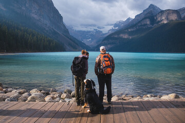 Young couple with backpacks and a dog. Tourists and travelers exploring Lake Louise in Banff, Canada. - obrazy, fototapety, plakaty