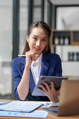 Asian business woman sitting happily with laptop and using tablet and taking intense notes on the work clipboard and smiling happily on the assignment.