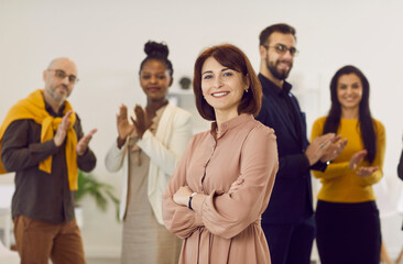 Portrait of successful female business professional and team leader. Happy, cheerful, beautiful woman in dress standing in office, with employees applauding in background. Success, recognition concept