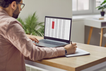 Concentrated male financier using laptop working in office with data entered in spreadsheet. Man fills out paper documents and enters data into electronic files marked in red on laptop screen. - obrazy, fototapety, plakaty