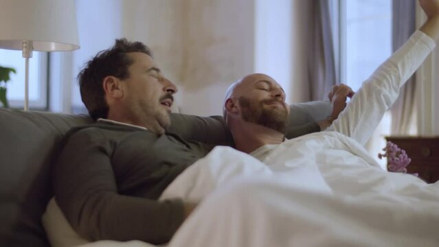 Happy homosexual men waking up in morning. Handsome bearded gay couple in pajamas lying in bed, hugging, yawning, stretching and talking after sound sleep at home. Love, LGBT, sleep cycle concept