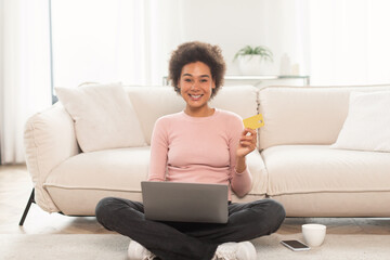 Fototapeta premium Cheerful young mixed race curly female shopaholic sit on floor with laptop, showing credit card, enjoy huge sale