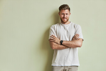 Minimal portrait of bearded young man smiling at camera while standing with arms crossed against pastel green wall, copy space