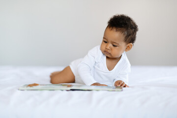 Naklejka premium Baby Development Activities. Cute Little Black Boy Relaxing In Bed With Book