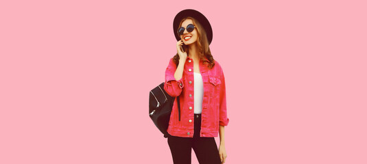 Portrait of happy smiling young woman calling on smartphone wearing jacket, black round hat, backpack on pink background