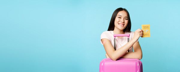Happy smiling asian girl, tourist stands with cute suitcase in airport, shows international...