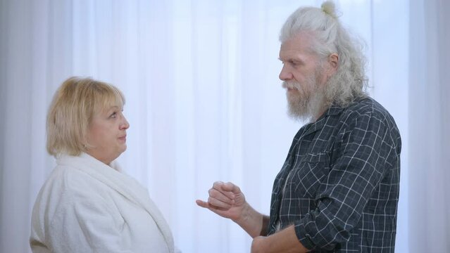Side view senior husband and wife making pinky swear shaking little fingers talking in slow motion. Caucasian man and woman making peace after quarrel standing in pajamas at home indoors