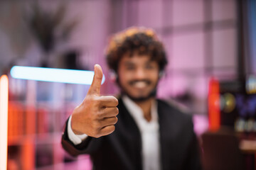 Focus on hand showing thumb up. Smiling positive confident man in formal wear and headset sitting at office desk. Company worker with beard looking at camera. Concept of people and business.