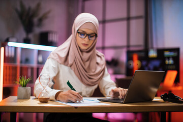 Fototapeta na wymiar Focus in hand with pen. Attractive confident muslim business woman, office manager, wearing hijab using laptop while making financial report while writing on paper working at night.