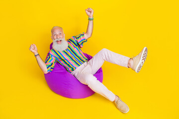 Full length photo of charming lucky man wear colorful shirt lying bean bag rising fists empty space isolated yellow color background