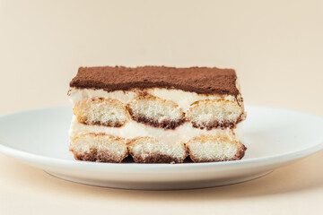Homemade tiramisu cake on white plate against beige background. Traditional Italian no-bake dessert made of savoiardi, filled with mascarpone cheese, coffee espresso and sprinkled with cocoa powder