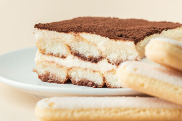 Homemade tiramisu cake on white plate against beige background. Traditional Italian no-bake dessert made of savoiardi, filled with mascarpone cheese, coffee espresso and sprinkled with cocoa powder
