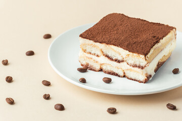 Homemade tiramisu cake on white plate against beige background surrounded by coffee beans . Italian dessert made of savoiardi cookies filled with mascarpone cheese and sprinkled with cocoa powder