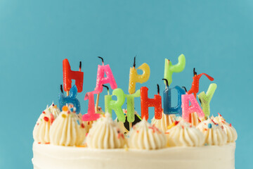 Birthday cake with white cream cheese frosting decorated with multicolored lit happy birthday text shaped candles on a blue background. Tradition of making a wish while blowing out candles on a cake