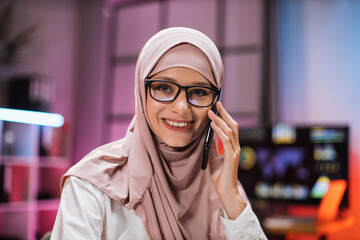 Close up portrait of attractive concentrated hardworking muslim businesswoman or manager in hijab having business conversation with smart phone in evening office. Concept of overwork