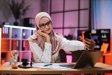 Portrait of smiling muslim businesswoman having mobile video conversation while sitting at table and working on wireless laptop. Working process at office of young girl at evening time.