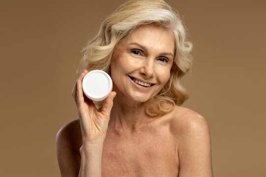 Isolated portrait on beige background of Caucasian attractive mature woman, smiling a beautiful toothy smile looking at camera, holding a jar with anti-aging smoothing cream near her face