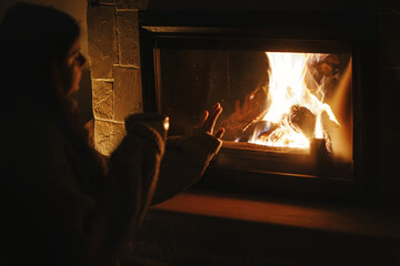 Woman holding cup of tea and warming up hands at cozy fireplace in dark evening room. Fireplace...