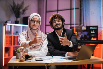 Confident two multi ethnic economists, young experienced muslim female and businessman, office manager in formal wear sitting at table using laptop in evening office showing thumb up.