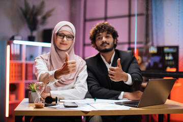 Confident two multi ethnic economists, young experienced muslim female and businessman, office manager in formal wear sitting at table using laptop in evening office showing thumb up.