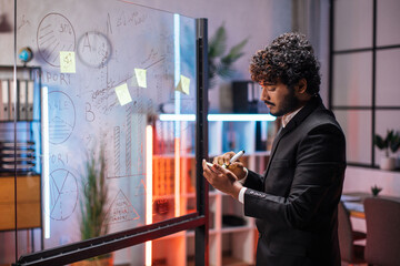 Focused male employee writing on colorful sticky notes on office glass board, developing business project, using tablet pc. Startup plan, people and technologies concept