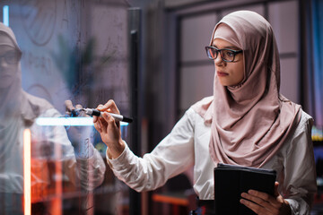 Side view of smart young businesswoman in hijab, brainstorming near office glass board, using...