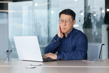 Fototapeta premium A young Asian man works in the office at a desk with a laptop. He holds his cheek. He feels a strong toothache, he needs the help of a dentist.