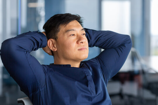 A Calm Young Asian Man Sits In The Office On A Chair, Leans Back, Hands Behind His Head, Looks Thoughtfully And Dreamily Out The Window. Resting From A Hard Day, Satisfied With Successful Work.