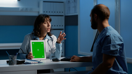 Medic holding tablet computer pointing at green screen chroma key display while discussing patient report with assistant. Clinical staff working night shift at medical expertise in hospital office