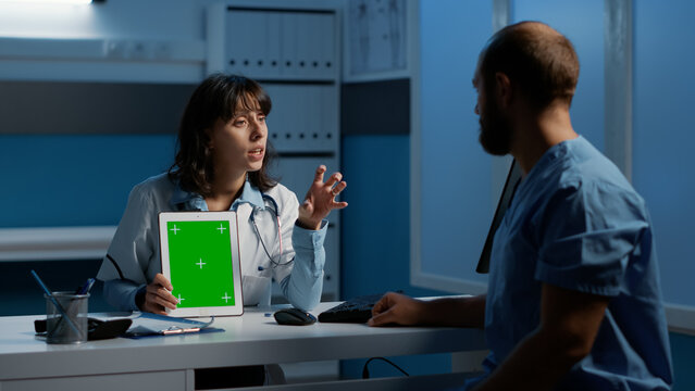 Medic Holding Tablet Computer Pointing At Green Screen Chroma Key Display While Discussing Patient Report With Assistant. Clinical Staff Working Night Shift At Medical Expertise In Hospital Office