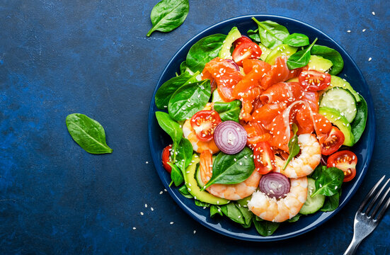 Salmon salad for ketogenic diet with shrimp, avocado, spinach, cucumber, tomato, cashew nuts, sesame. Low-carbohydrate lunch rich in healthy fats. Blue table background, top view
