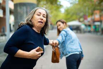 Street thief trying to steal handbag of scared elderly woman walking through city on warm autumn day