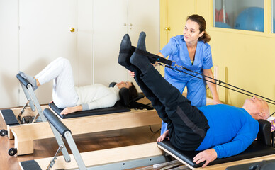 Fototapeta na wymiar Senior man practicing pilates system on reformer in rehabilitation center to improve and maintain mobility under supervision of qualified female doctor