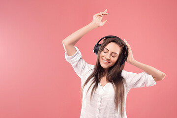Beautiful young woman in wireless headphones listening to music and dancing on pink background. Girl uses wireless earphones