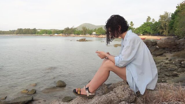Caucasian woman is sitting on stone in front of sea and texting message on smartphone in her hands. Female holiday maker is resting on the shoreline in warm country during vacations and using mobile
