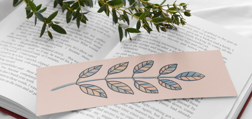 Open book with beautiful bookmark and green branches on bed, closeup