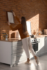 Young woman hanging blank frame on brick wall in kitchen