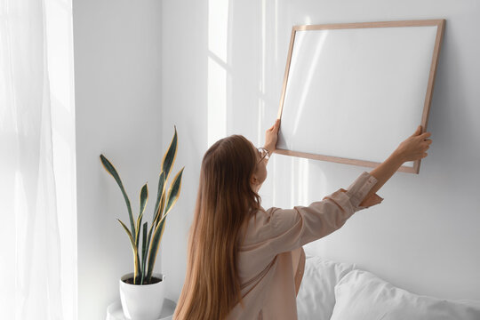 Young woman hanging blank frame on light wall in bedroom
